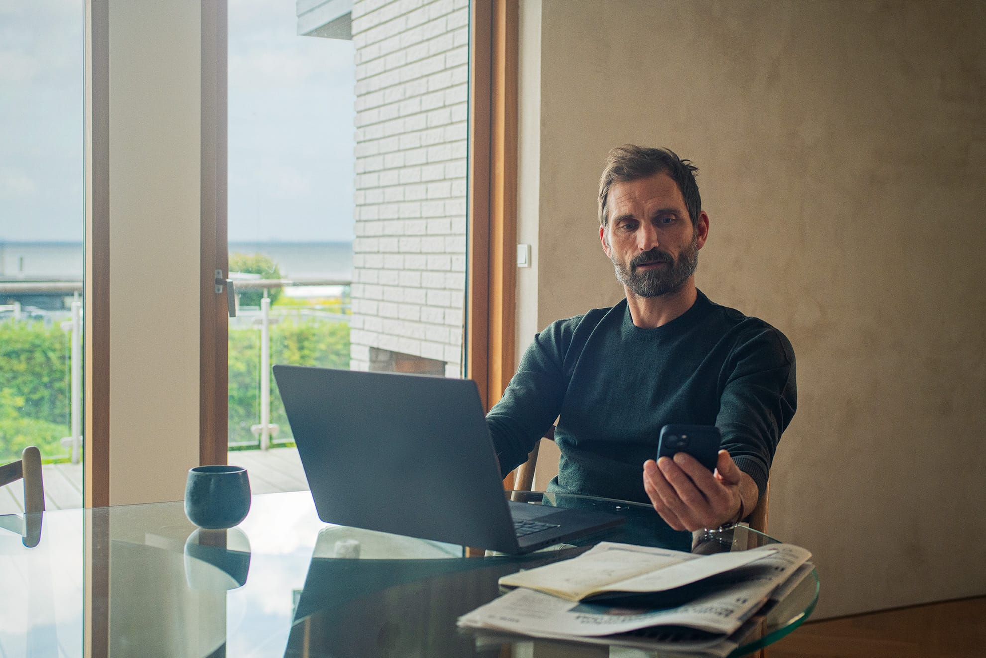 En mand sidder ved et bord med en bærbar computer og en smartphone i hånden. Han arbejder i et lyst rum med papirer og en kop kaffe.