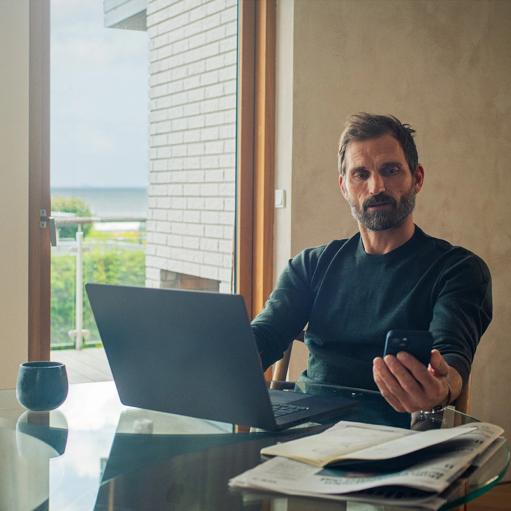 En mand sidder ved et bord med en bærbar computer og en smartphone i hånden. Han arbejder i et lyst rum med papirer og en kop kaffe.