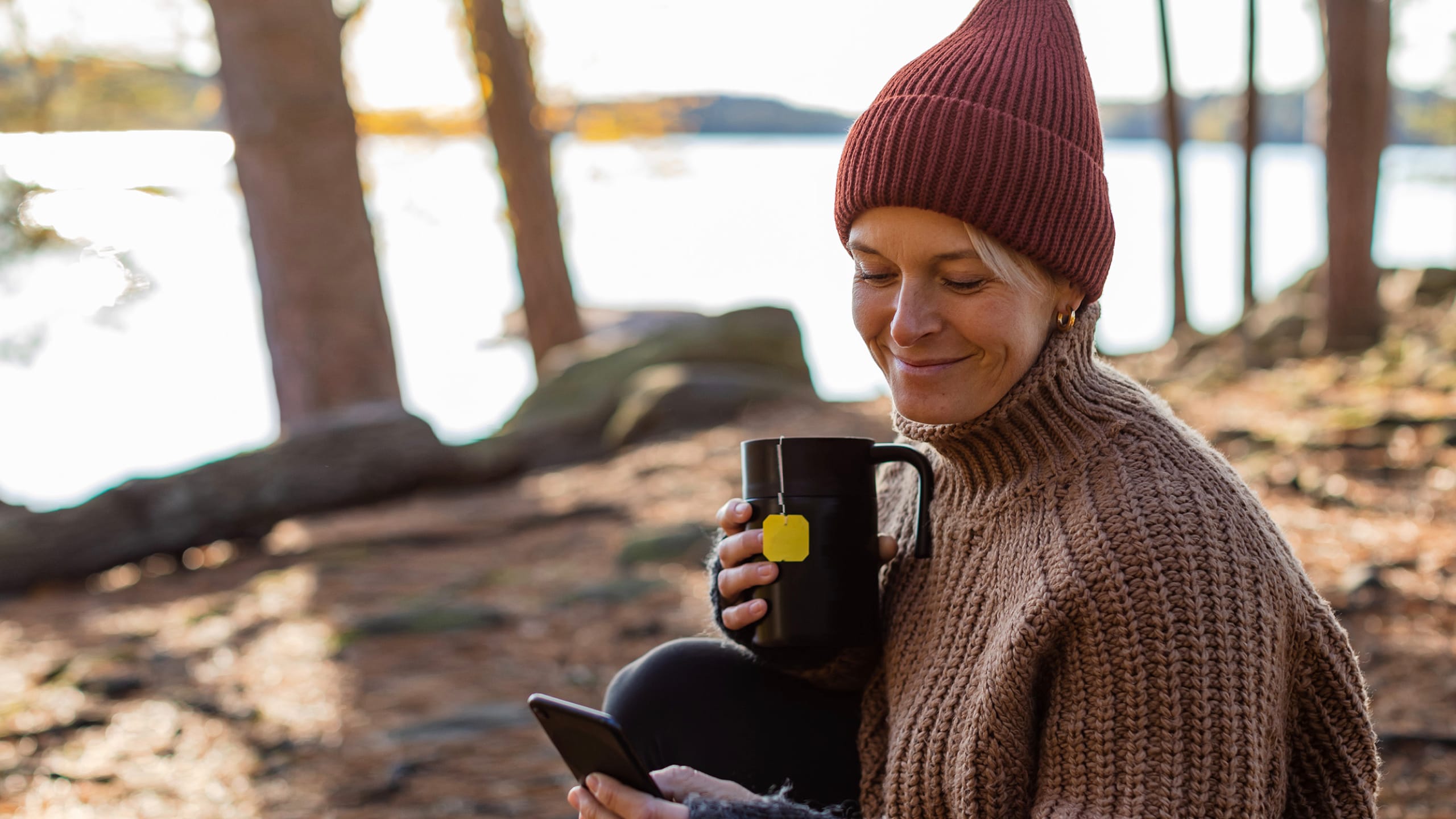 Kvinde i en skov med en kop kaffe, kigger på en mobiltelefon