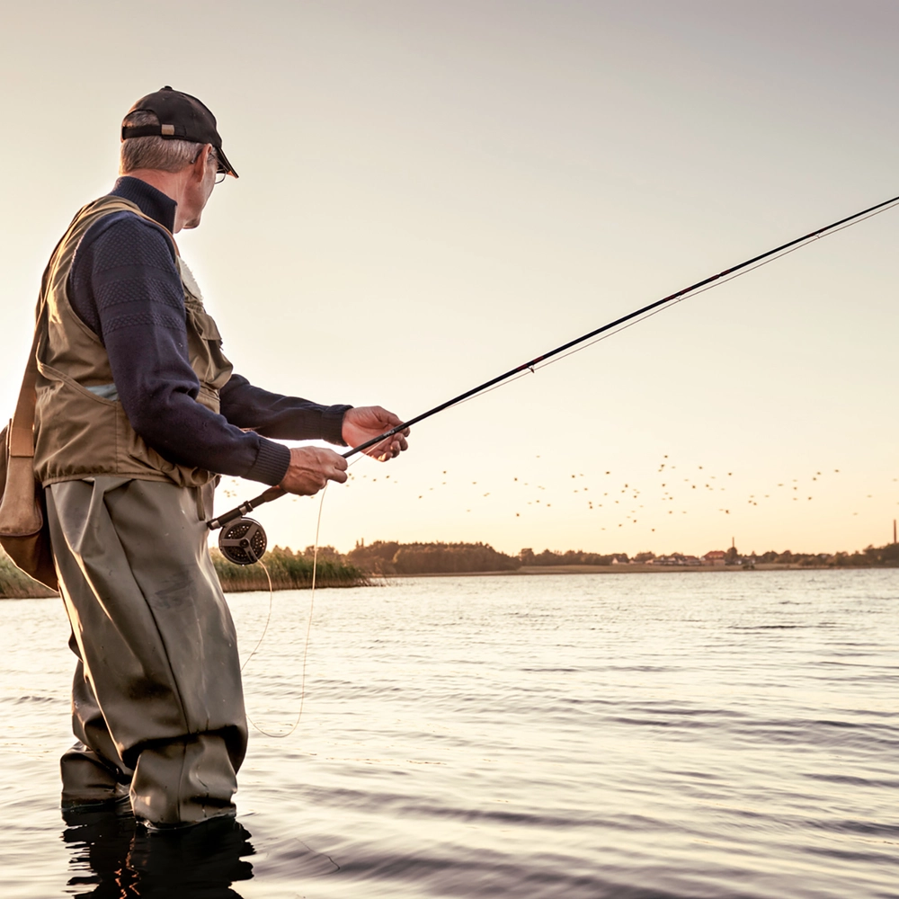 Mand iført waders står ude i vandet og fisker med fluestang