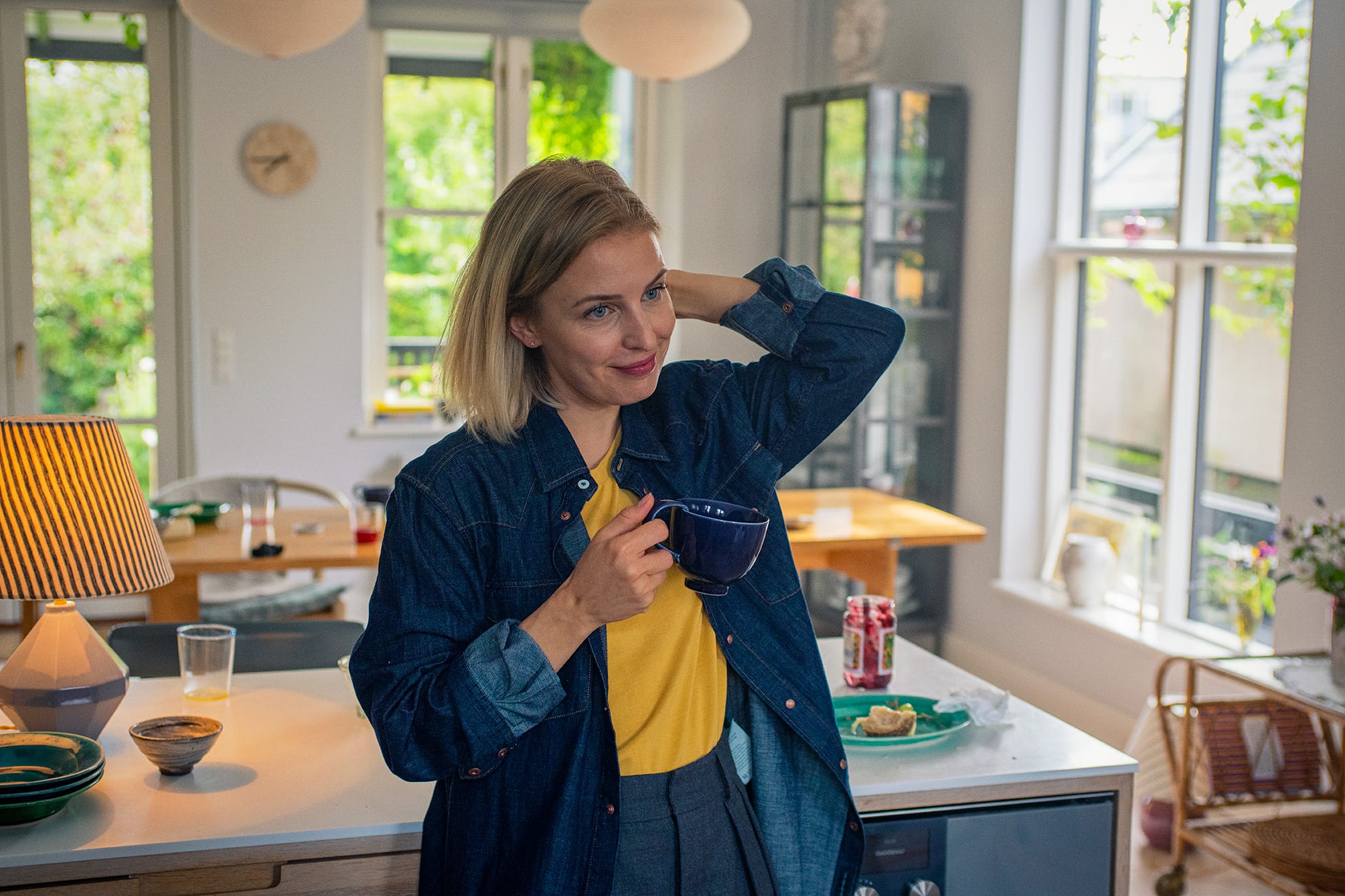 En kvinde står i et lyst køkken med en kop i hånden og smiler. Hun har blondt hår, en gul bluse og en åben denimskjorte. I baggrunden ses et spisebord med mad og glas samt store vinduer med udsigt til grønne træer.