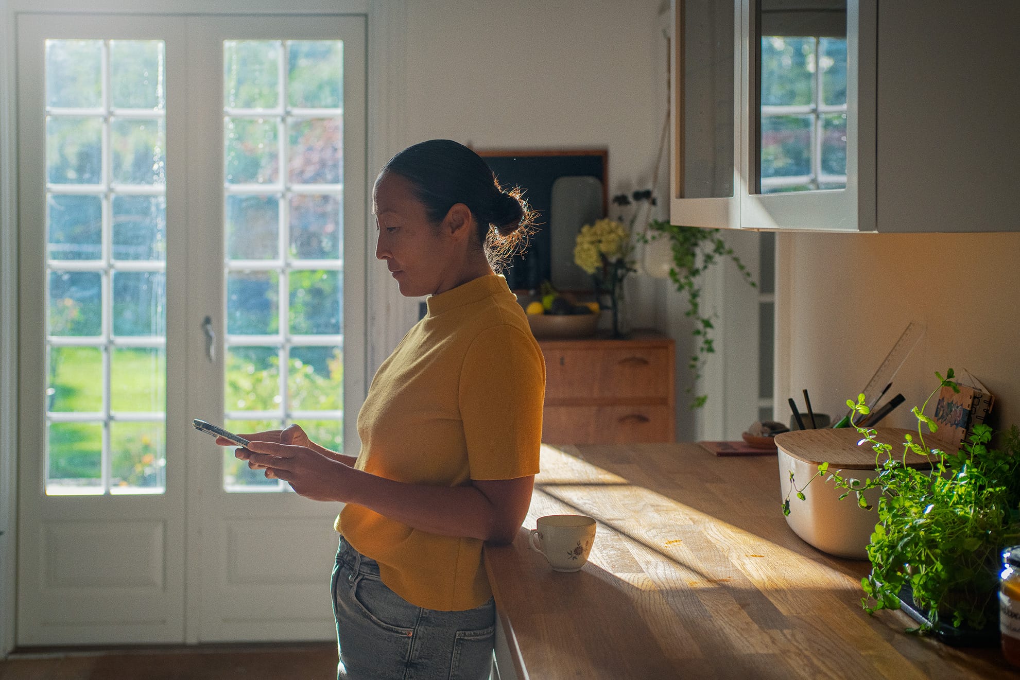 En kvinde står i et solbelyst køkken og kigger på sin telefon. Hun har en gul bluse på og læner sig op ad køkkenbordet, hvor der står en kop. Bag hende ses en glasdør med udsigt til en grøn have.