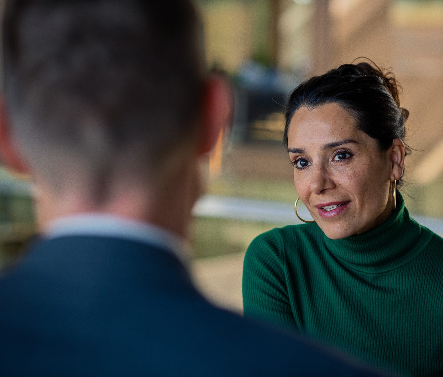 En kvinde i grøn rullekrave taler med en anden person i et professionelt miljø. Hun ser engageret ud. Baggrunden er sløret og viser et lyst kontorområde.